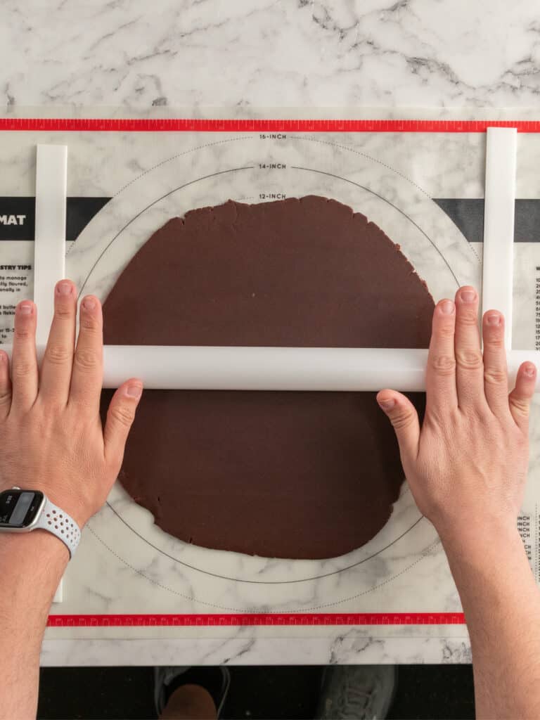 Hands rolling out chocolate dough with a rolling pin on a pastry mat with measurement markings, on a marble countertop.
