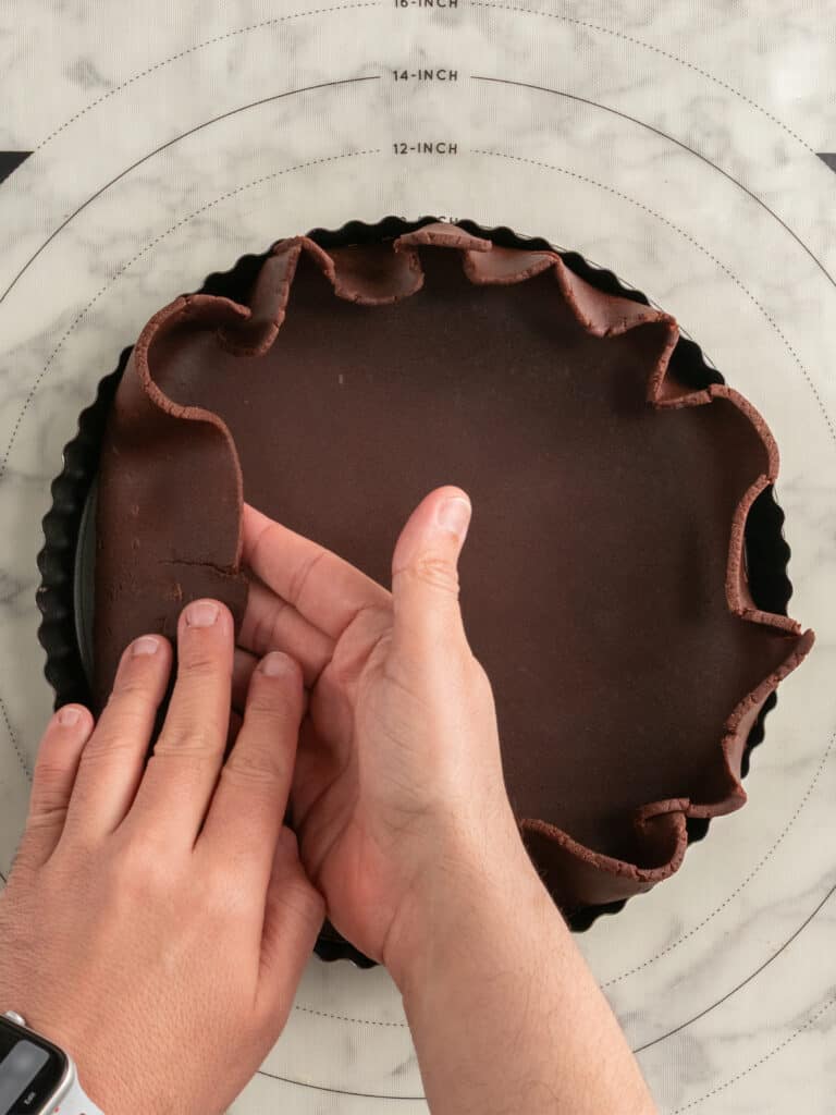 Hands gently press chocolate pie dough into a round tart pan on a marble surface, with excess dough draping over the pans edges. A watch is visible on one wrist.
