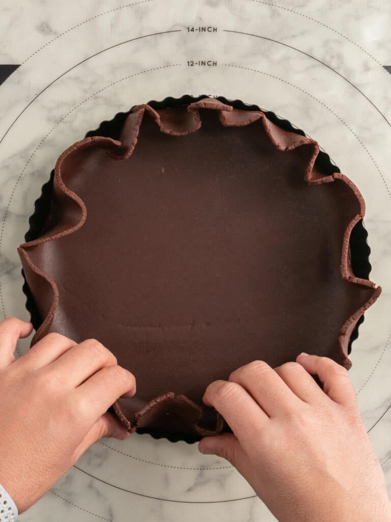 Two hands press chocolate dough into a round tart pan on a marble surface with a measurement guide in the background.