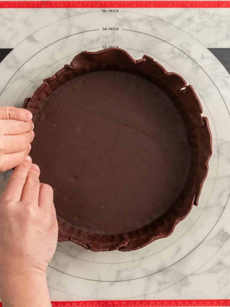 Hands pressing chocolate pie crust dough into a round tart pan on a marble surface with measurement markings.