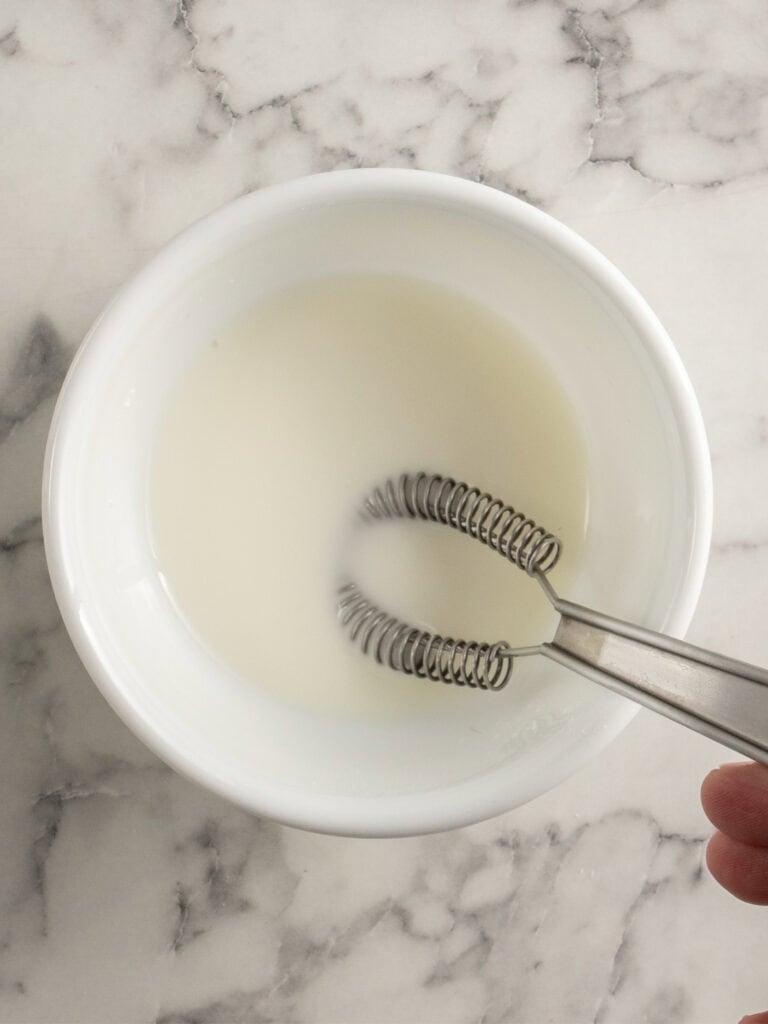 A hand whisking white liquid in a white bowl on a marble countertop.