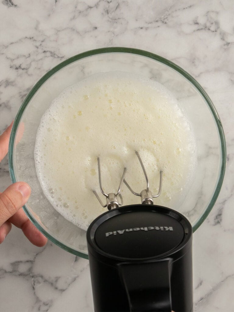 A hand holds a glass bowl with frothy egg whites being whisked by an electric hand mixer on a marble countertop.