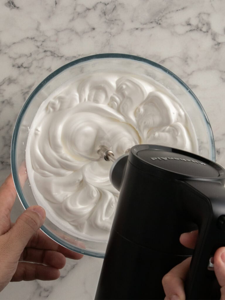 A person uses a black electric hand mixer to whip egg whites in a glass bowl on a marble countertop.
