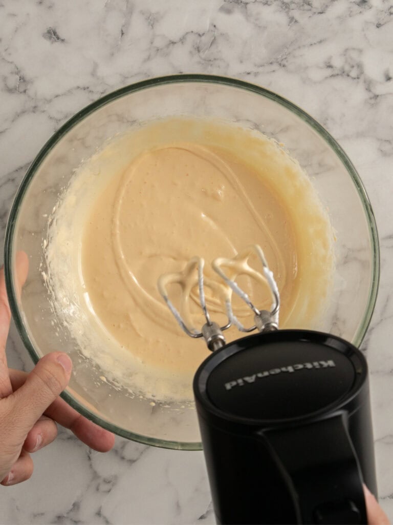 A hand holds a glass bowl with creamy batter while using a black electric hand mixer, seen from above on a marble countertop.
