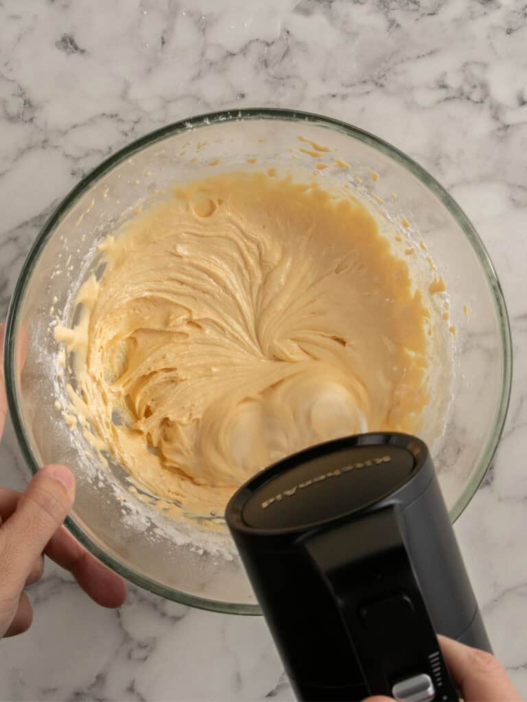 A hand holds an electric mixer blending creamy batter in a glass bowl on a marble countertop.