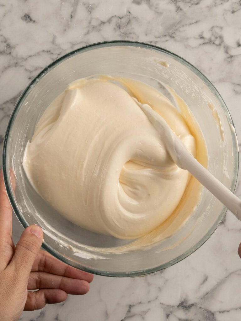 A hand holds a glass bowl of thick, creamy batter being gently mixed with a white spatula on a marble countertop.