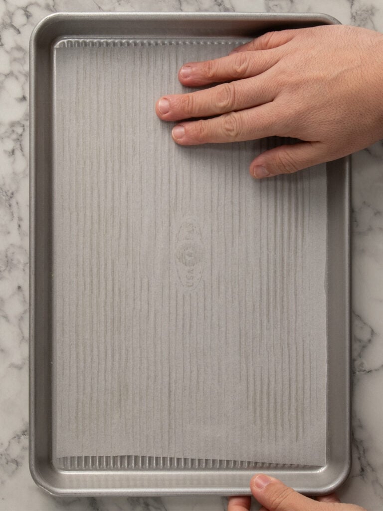 A hand presses a sheet of parchment paper onto a metal baking tray, which rests on a white marble surface.