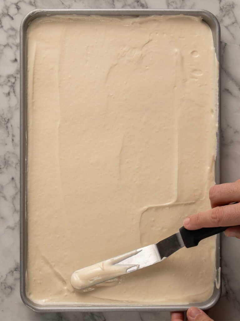 A hand uses an offset spatula to spread cake batter evenly in a metal baking pan on a marble countertop.