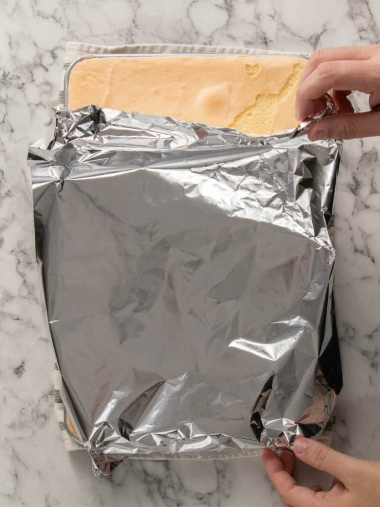 A person lifts a sheet of aluminum foil covering a rectangular sponge cake on a marble countertop.