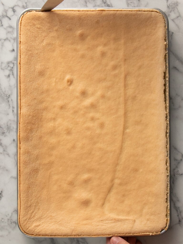 A rectangular sheet cake with a golden-brown top sits on a marble surface. A hand holds the pan, while a knife is inserted along the cakes edge to loosen it.