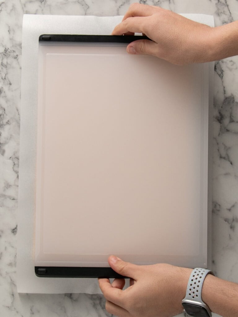 Two hands place a rectangular baking sheet upside down on a sheet of parchment paper on a marble countertop. One wrist wears a gray watch with perforated holes.