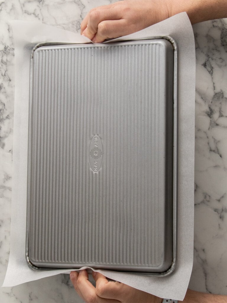 Two hands hold a baking sheet upside down on a piece of parchment paper on a marble countertop.
