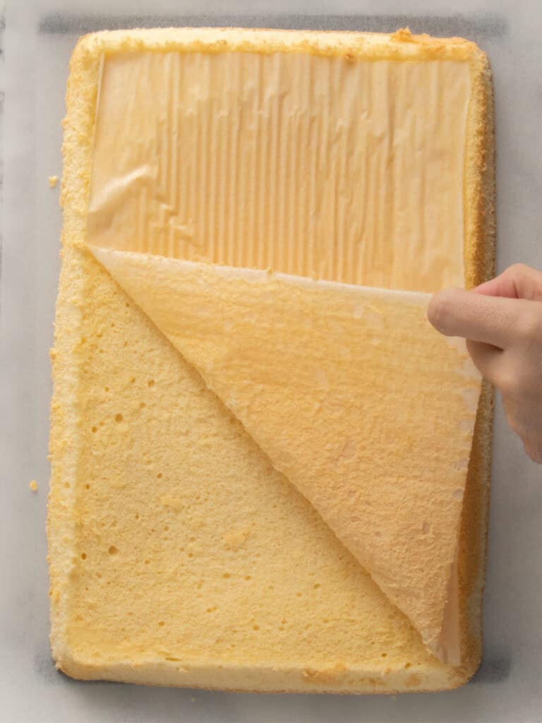 A hand peels parchment paper from a rectangular, freshly baked sponge cake resting on a sheet of baking paper.