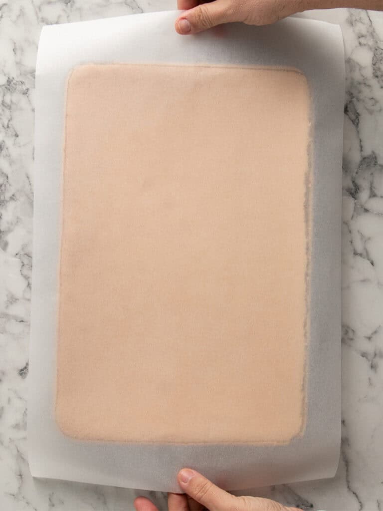 Two hands hold a rectangular baking pan lined with parchment paper and containing a thin, evenly-baked, light brown sheet cake, placed on a white marble surface.