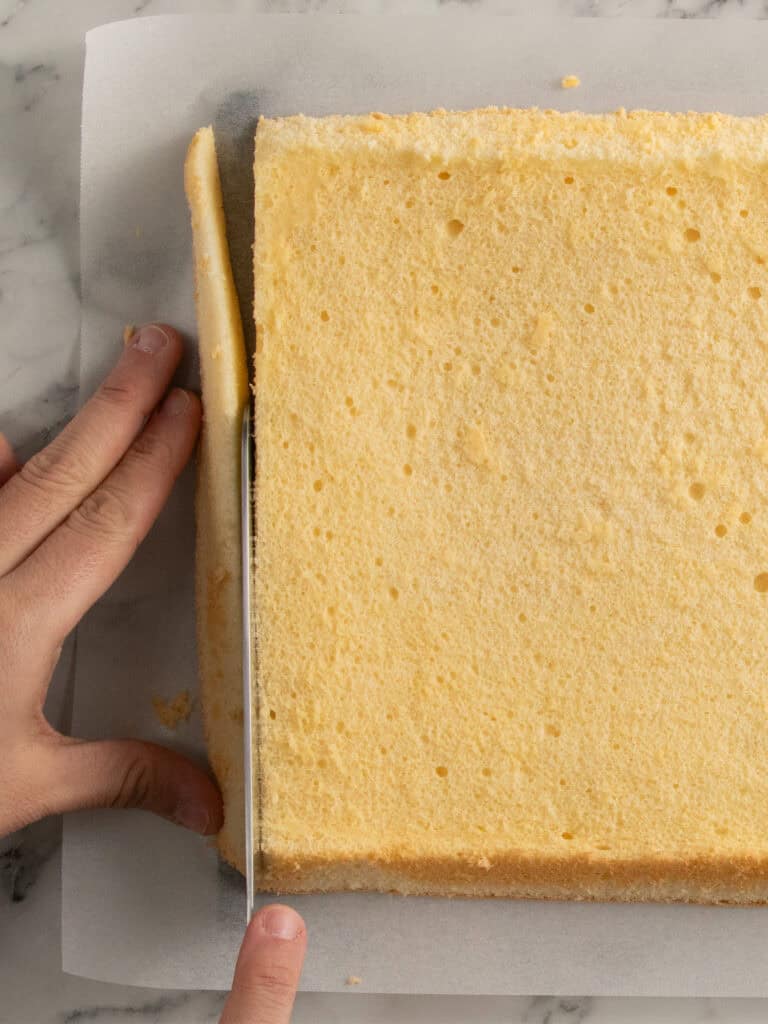 A hand uses a knife to trim the edge of a rectangular sponge cake on parchment paper, placed on a marble surface.