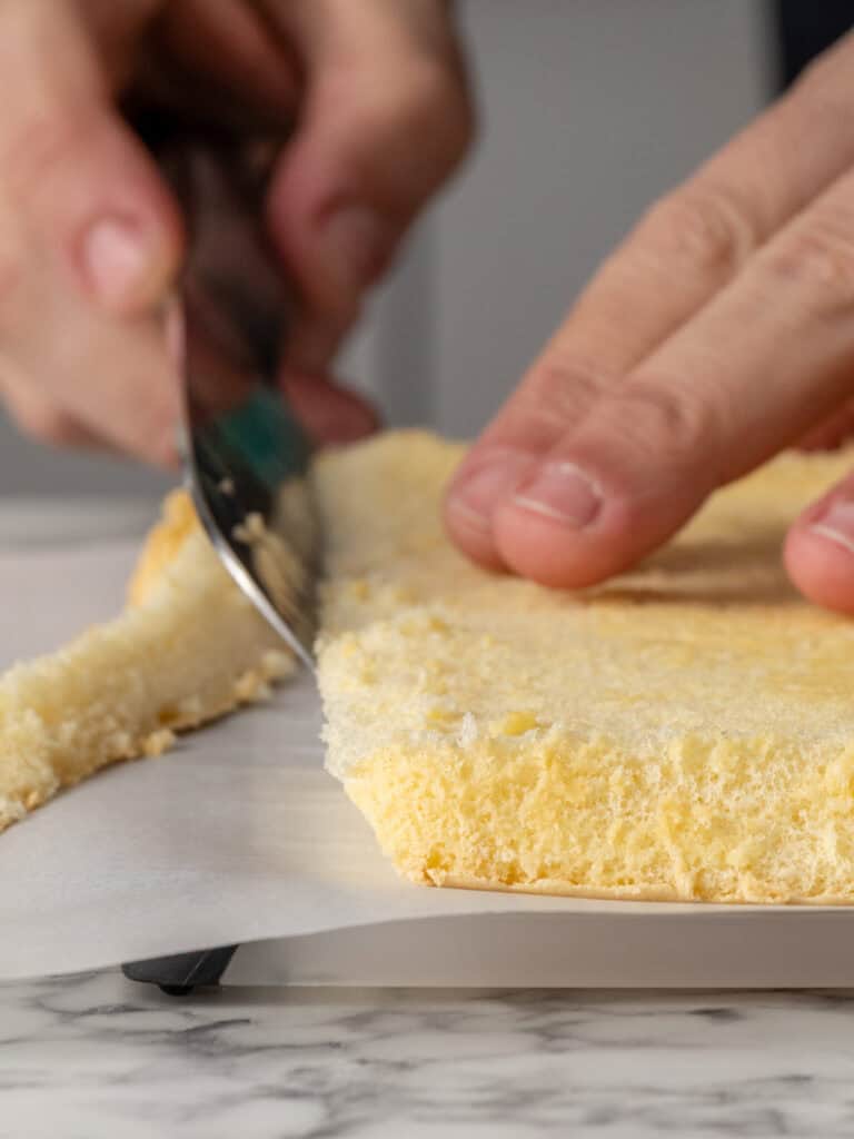 A close-up of hands using a knife to trim the edge of a round, yellow sponge cake on parchment paper, with a marble countertop underneath.