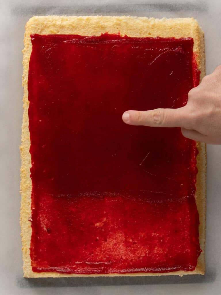 A hand points to a rectangular sponge cake topped with a smooth layer of red jam, placed on a light-colored surface.