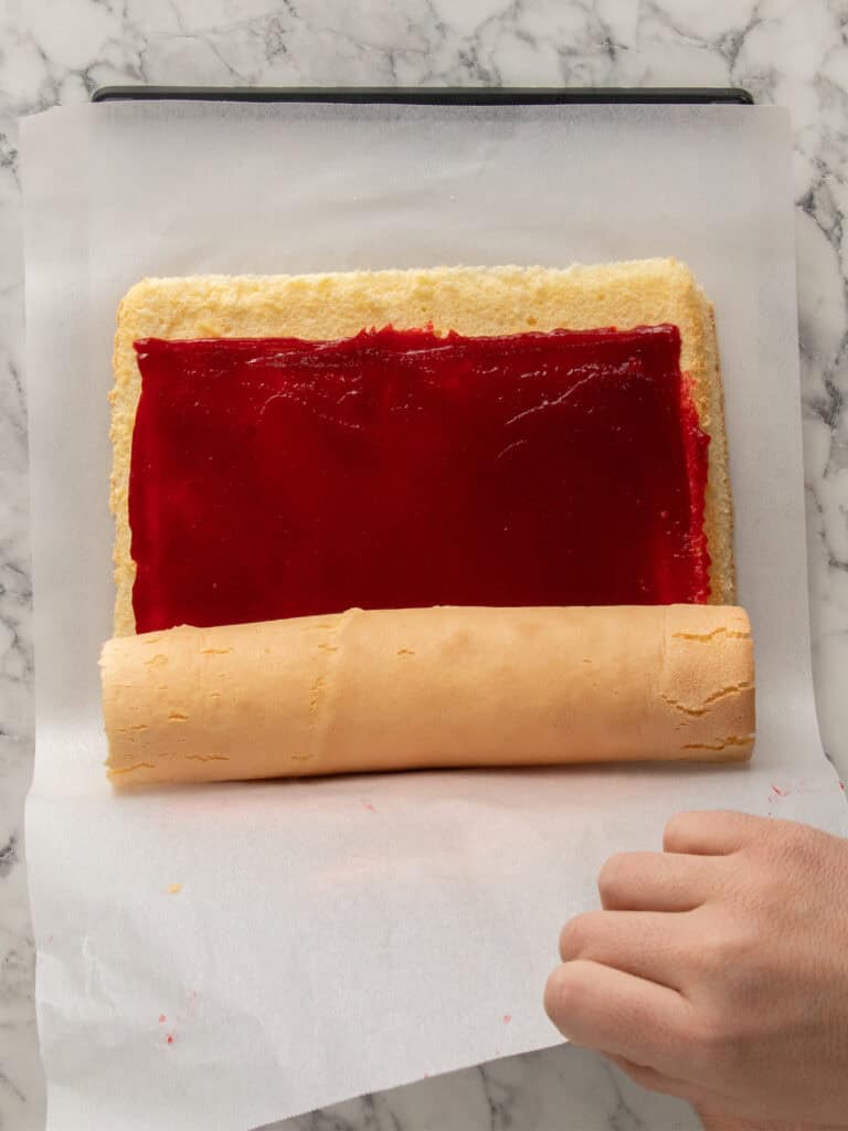 A hand rolls a sponge cake spread with red jam on parchment paper over a marble surface.