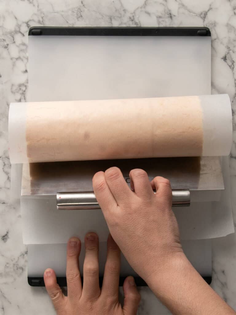 A hand uses a metal scraper to flatten dough between sheets of parchment paper on a marble surface.