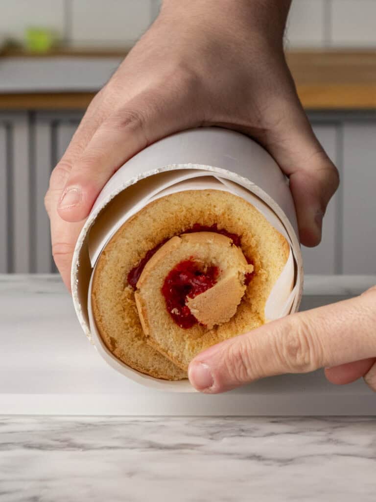 Close-up of hands holding a rolled sponge cake with jam filling, partially wrapped in parchment paper on a marble surface.