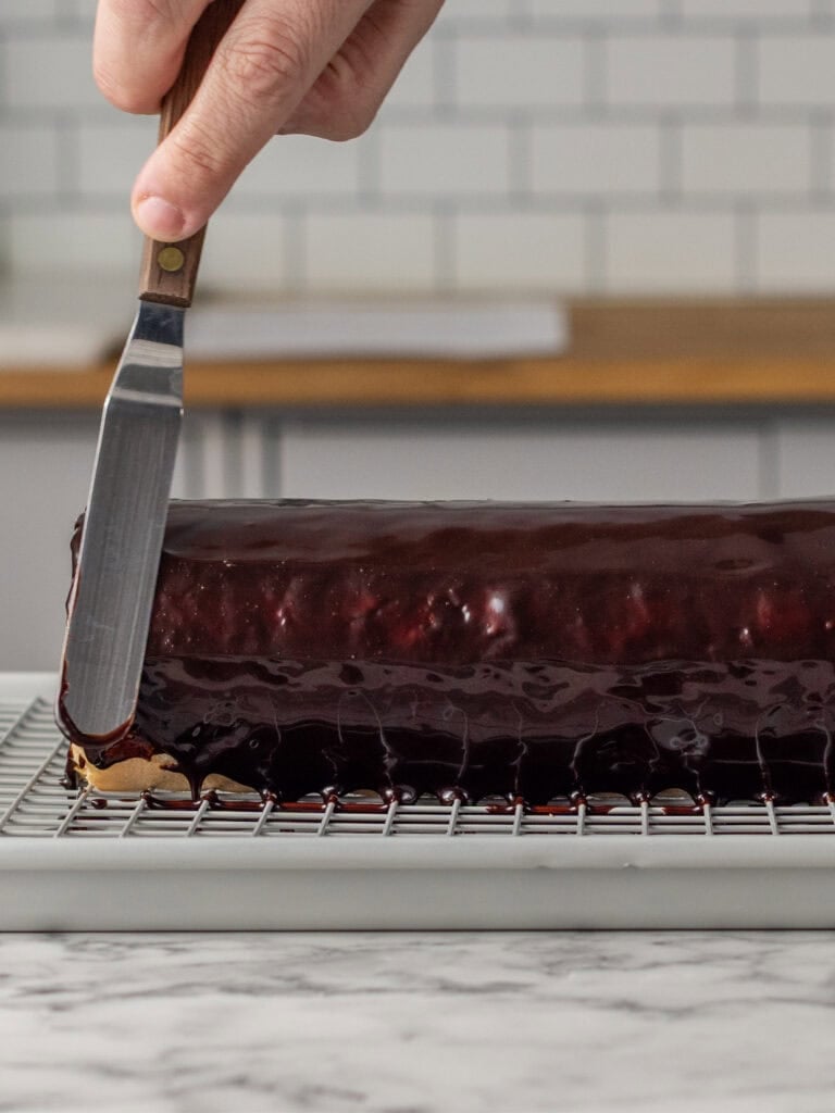 A hand uses an offset spatula to smooth glossy chocolate glaze over a rectangular cake on a cooling rack, with a kitchen counter in the background.