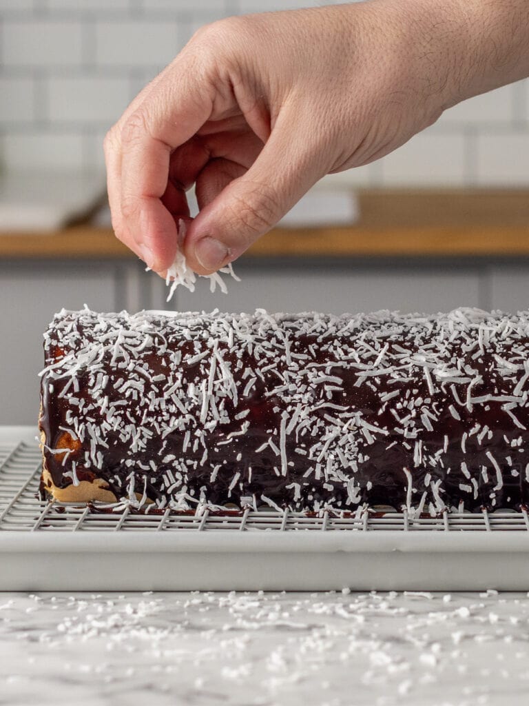A hand sprinkles shredded coconut onto a rectangular chocolate-covered cake resting on a wire rack, with a marble counter and kitchen in the background.