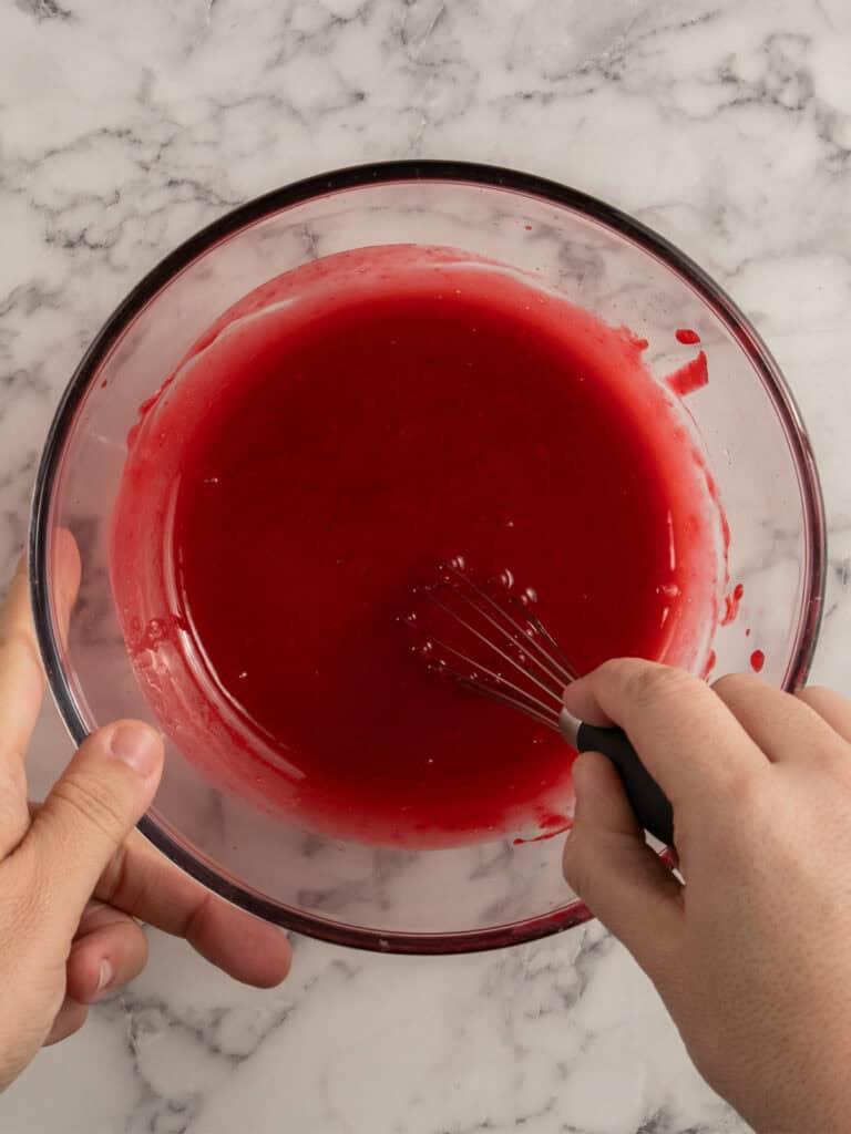 A person whisking a bowl of vibrant red liquid on a marble countertop.