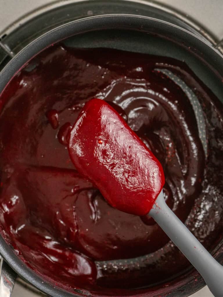 A close-up of a saucepan filled with smooth, glossy chocolate ganache being stirred with a gray spatula.
