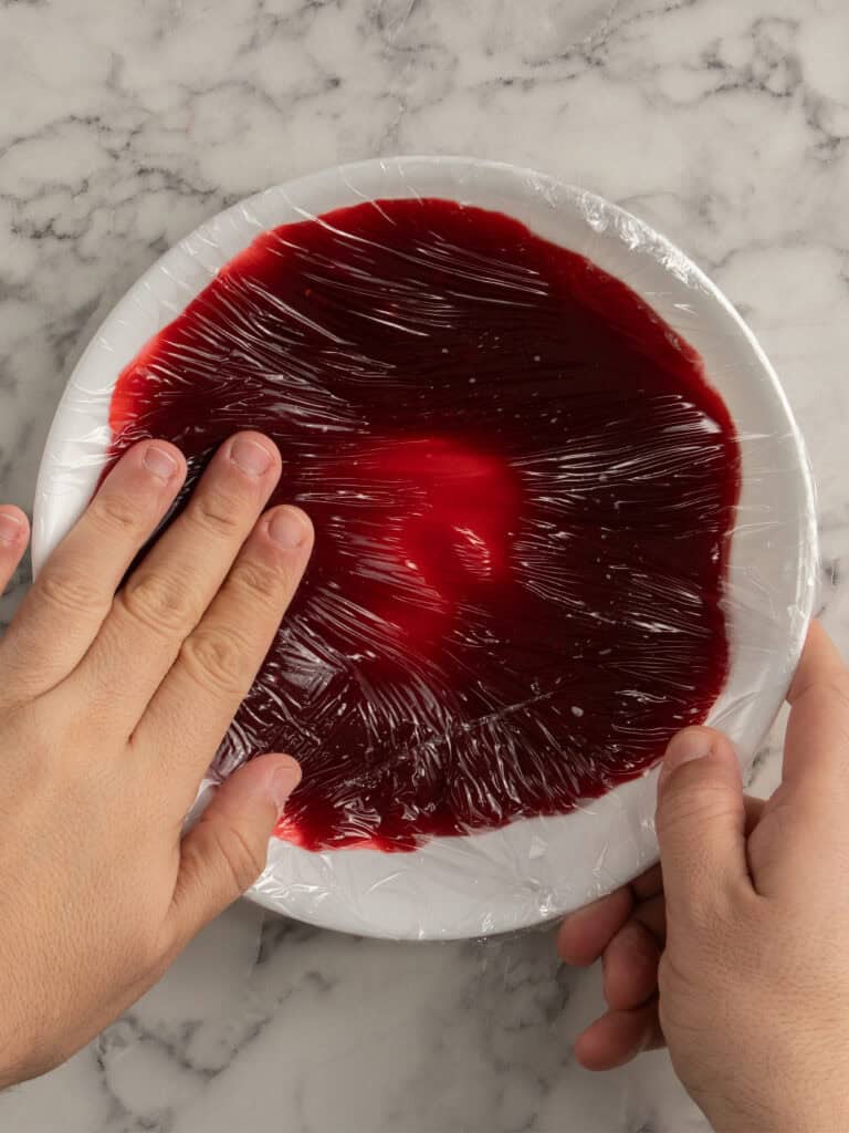 Red jelly in a white plate covered with plastic wrap on marble surface.