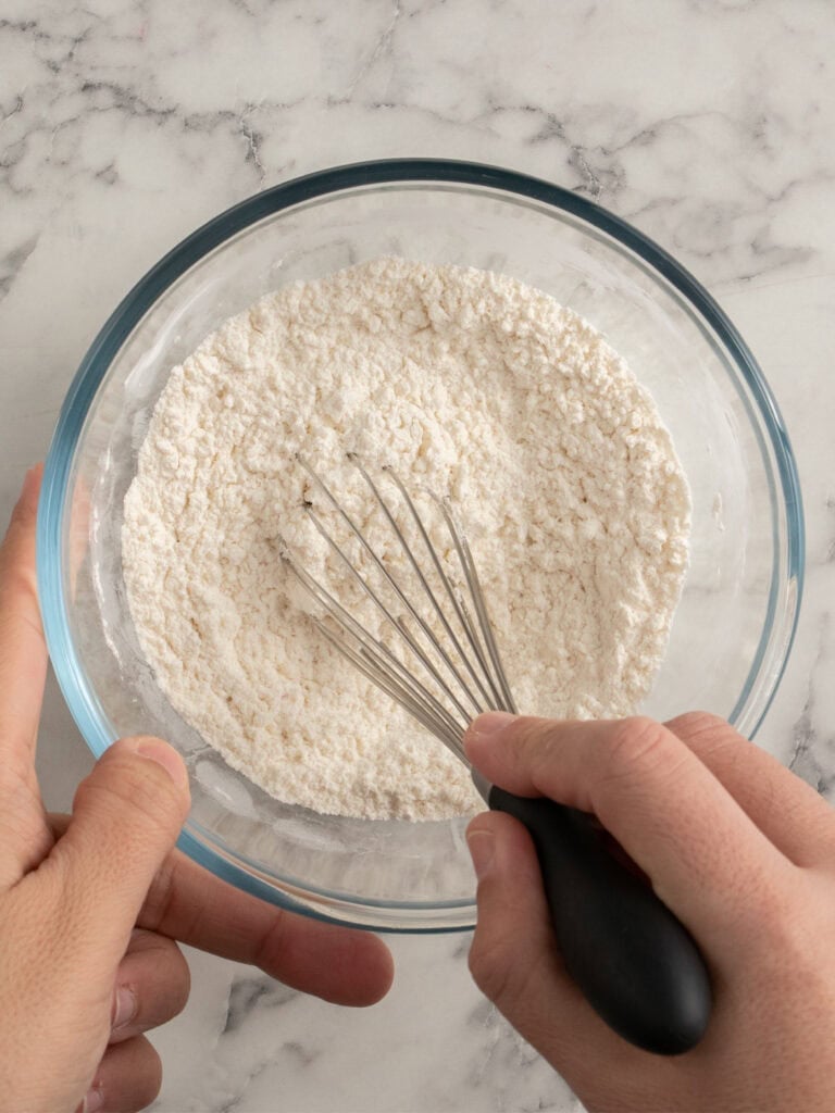 A person uses a whisk to mix flour in a clear glass bowl on a marble countertop.