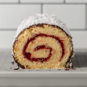 A close-up of a sliced jelly roll cake with a spiral of red filling, coated in coconut and chocolate on the outside, resting on a wire rack in front of a white tiled background.