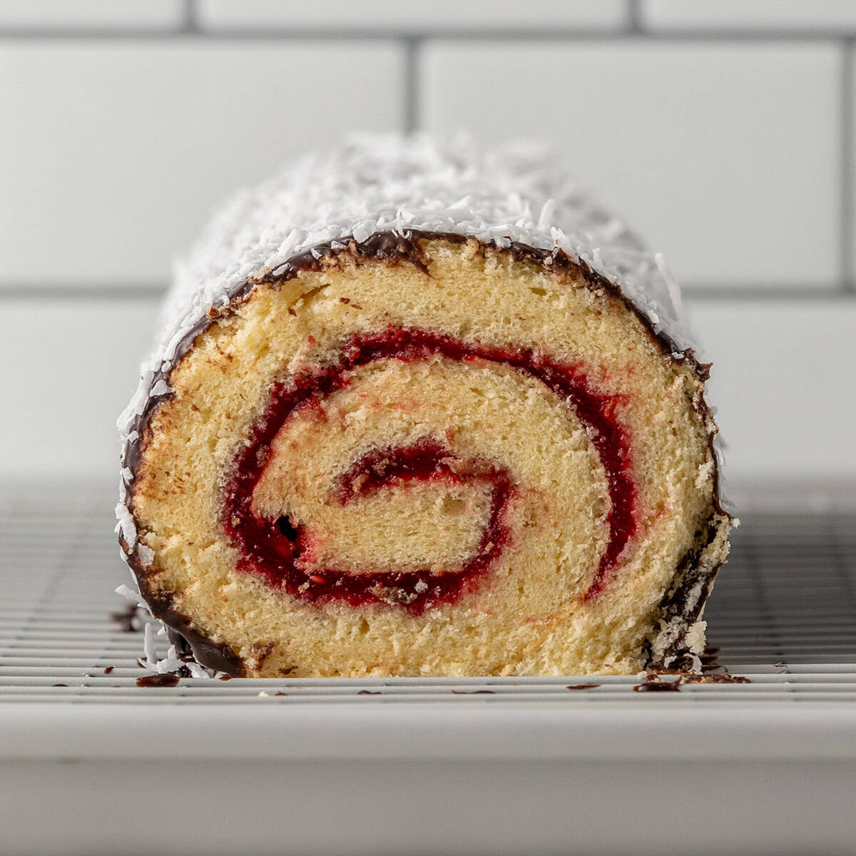 A close-up of a sliced jelly roll cake with a spiral of red filling, coated in coconut and chocolate on the outside, resting on a wire rack in front of a white tiled background.