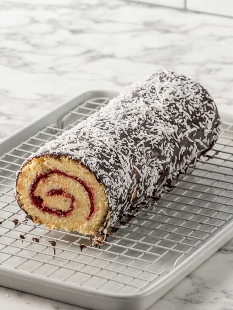 A chocolate-covered rolled sponge cake with a raspberry swirl filling, topped with shredded coconut, sits on a wire cooling rack over a baking tray on a marble countertop.
