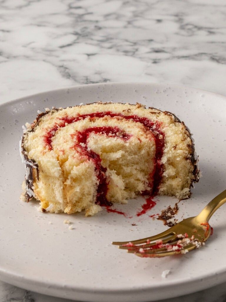 A slice of Swiss roll cake with raspberry filling sits on a white plate. A gold fork rests nearby, and crumbs are scattered around. The background is a white marble surface.