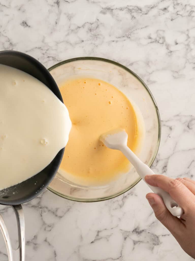 A hand stirs a bowl of beaten eggs while hot milk is being poured in from a saucepan on a marble countertop.