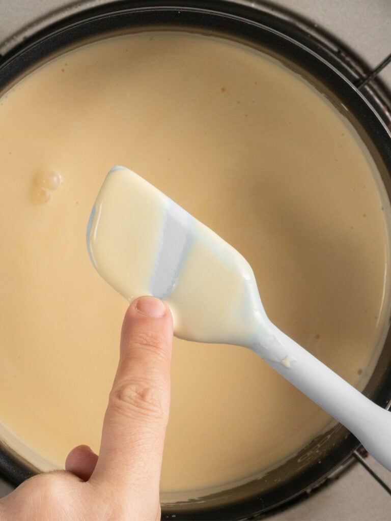 A hand runs a finger across a spatula coated in a thick, creamy sauce held over a saucepan filled with the same sauce.