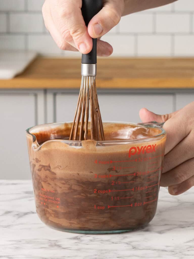 A person uses a whisk to mix chocolate batter in a glass Pyrex measuring cup on a marble countertop.