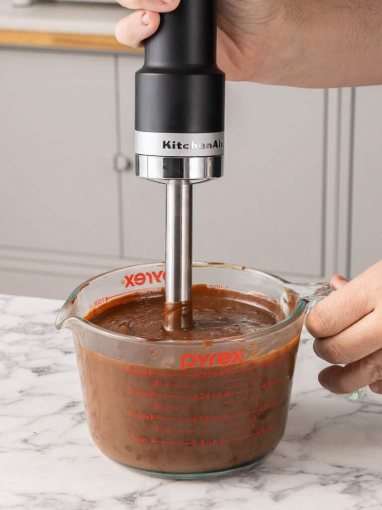 A person uses an immersion blender to mix chocolate batter in a glass measuring cup on a marble countertop.