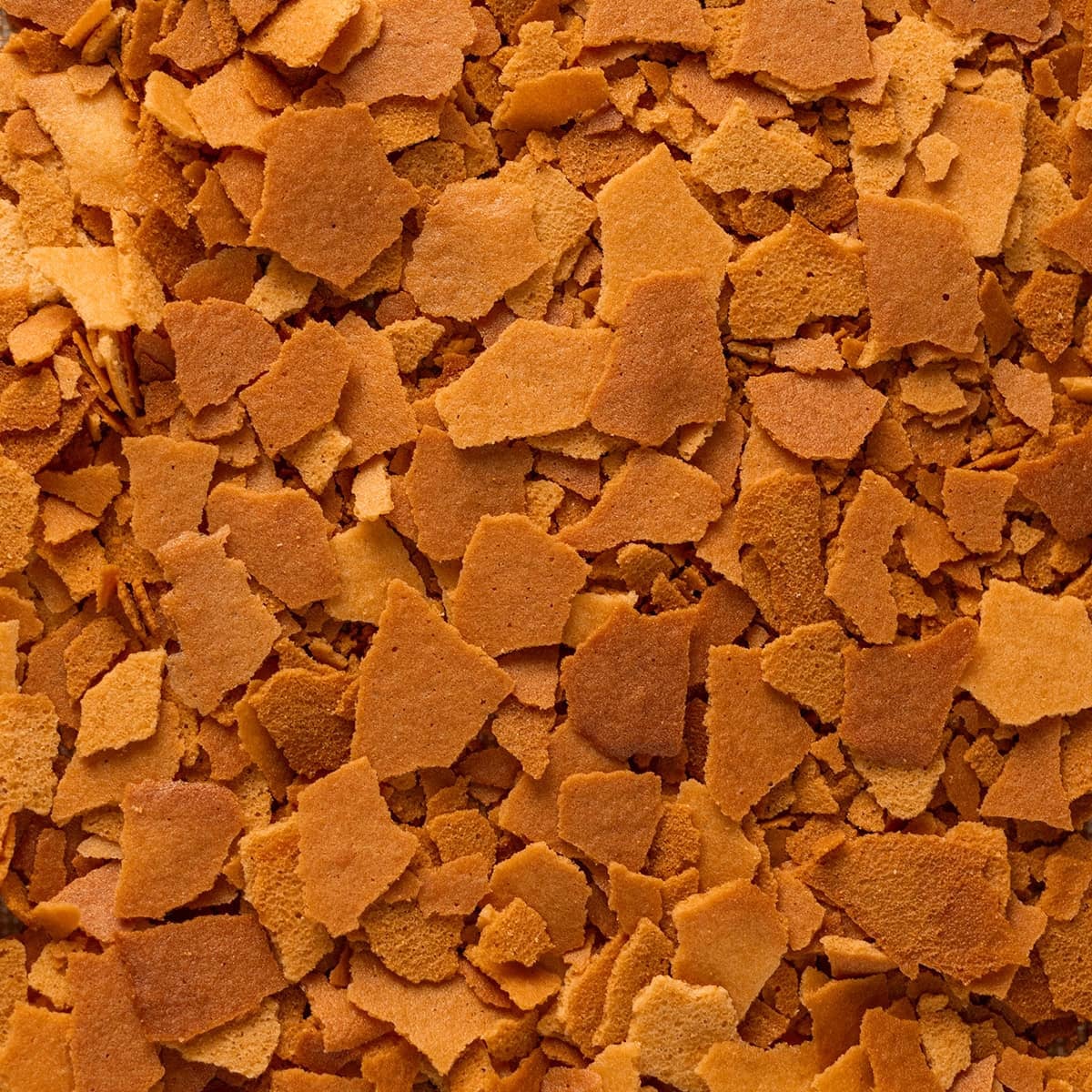 Close-up of scattered golden-brown flakes, resembling baked cereal or pastry pieces, creating a textured surface.