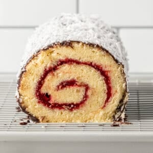 A close-up of a rolled sponge cake with a red jam swirl, coated in chocolate and shredded coconut, sitting on a wire cooling rack.