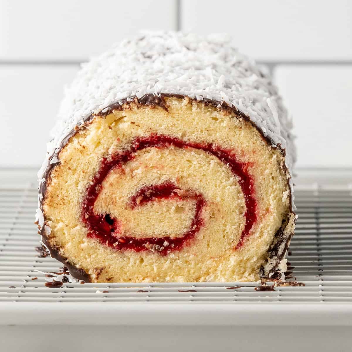 A close-up of a rolled sponge cake with a red jam swirl, coated in chocolate and shredded coconut, sitting on a wire cooling rack.