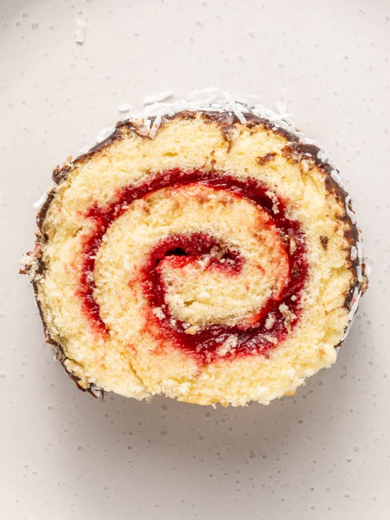 A close-up of a slice of jam roll cake with a visible red jam swirl, light yellow sponge, chocolate coating on the edges, and a sprinkle of shredded coconut on top, placed on a light-colored surface.