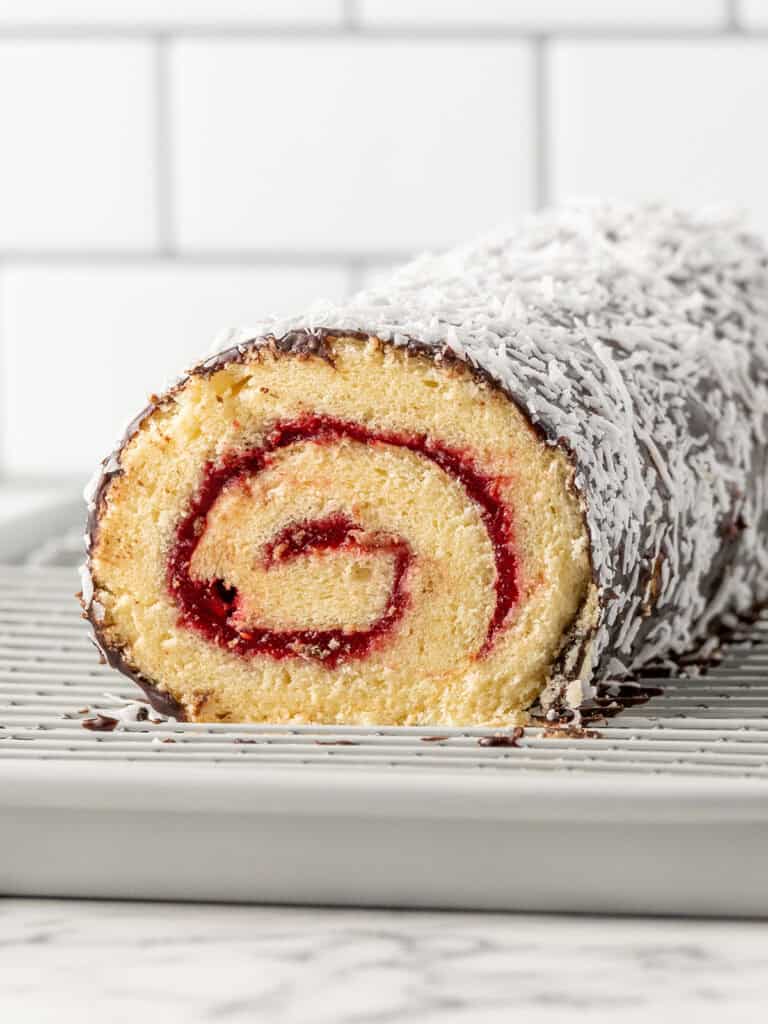 A rolled sponge cake with a red jam swirl, coated in chocolate and shredded coconut, sits on a metal cooling rack against a white tile background.