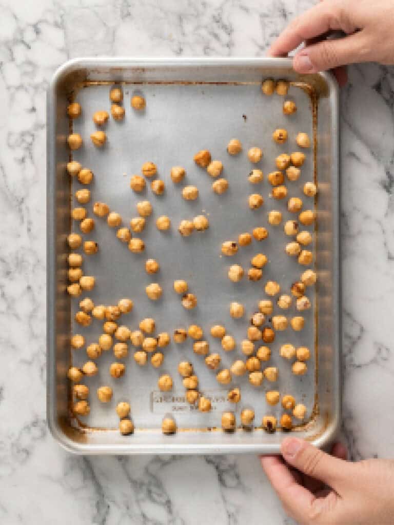 Hands holding a metal baking sheet filled with toasted hazelnuts on a marble surface.
