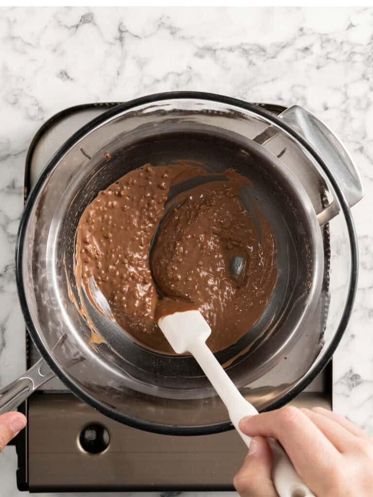 A hand uses a spatula to stir melting chocolate with rice crispies in a glass bowl over a stovetop.