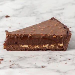 A slice of chocolate tart with a nut-filled base on a marble surface.