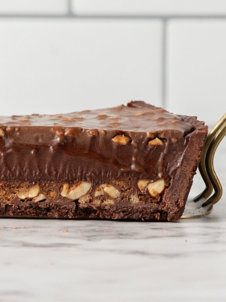 A slice of chocolate nut tart with a dark crust, lifted by a gold cake server.
