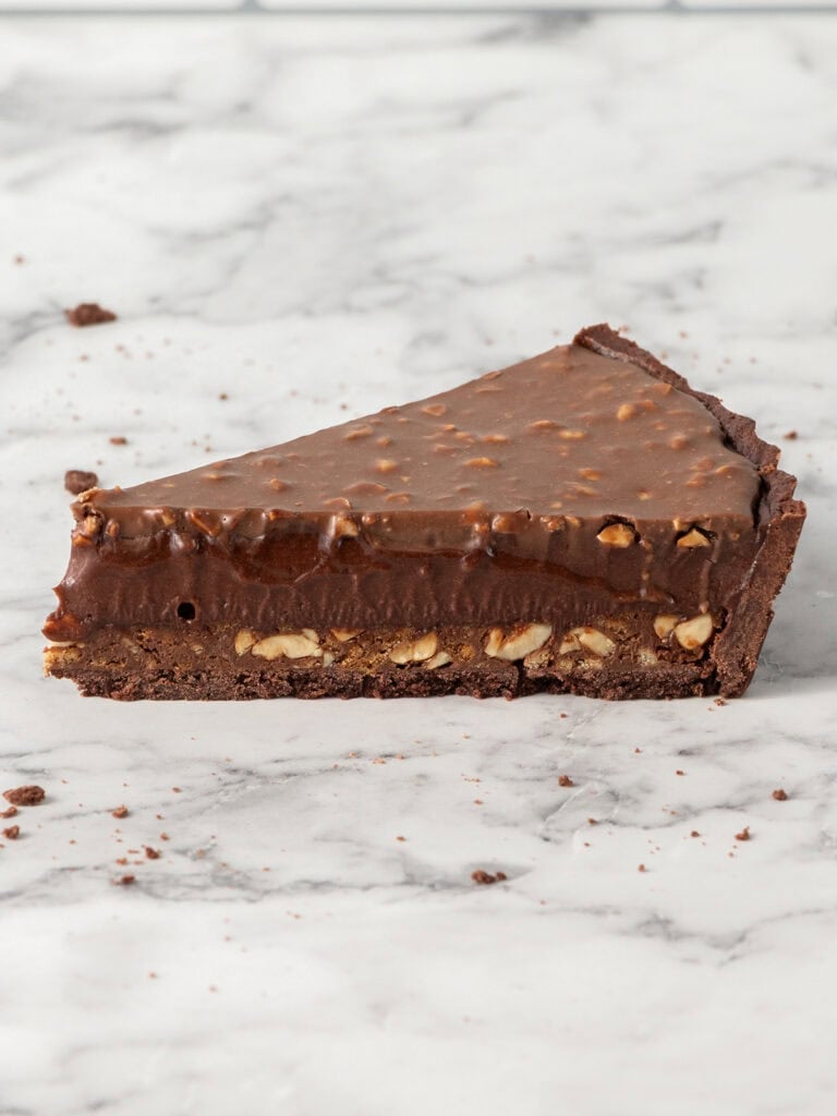 A slice of chocolate nut tart with a crunchy base, a dark chocolate filling, and a smooth chocolate ganache topping on a marble surface.
