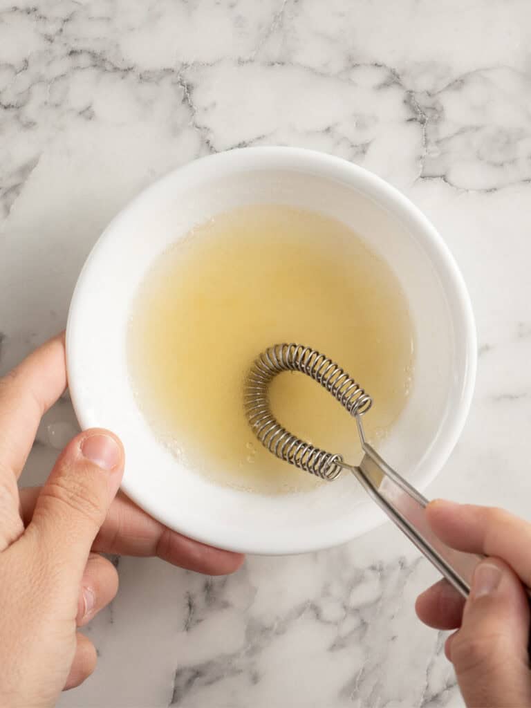 A metal coil whisk mixing a pale yellow liquid in a white bowl.