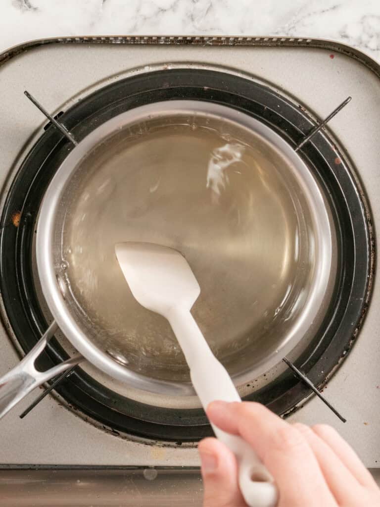 A hand uses a white spatula to stir a clear liquid in a saucepan on a stovetop.